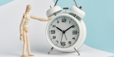 Wooden man stands with his hand on the alarm clock. Time management, waiting, punctuality.